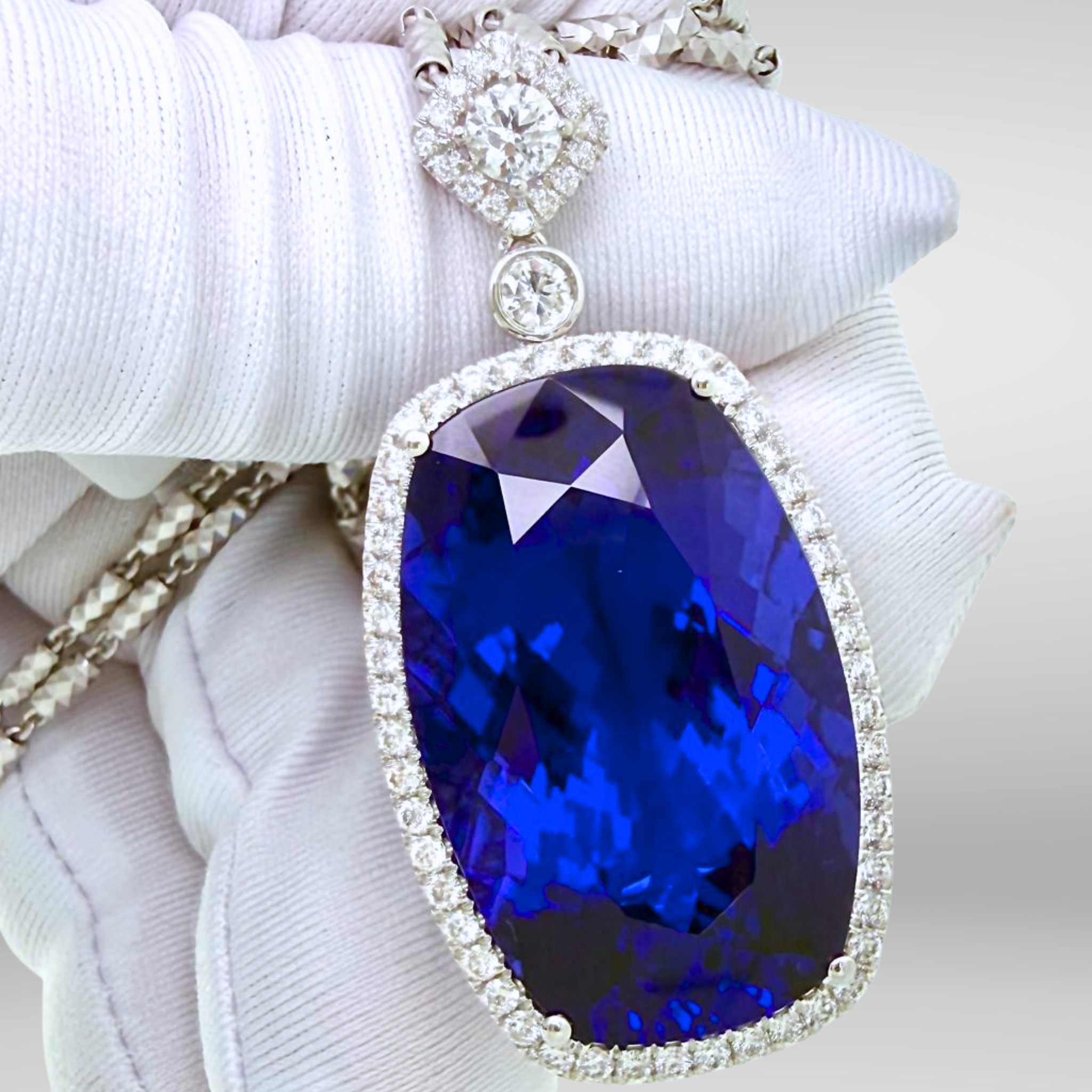 A close-up of a white-gloved hand holding a large oval tanzanite investment grade 50 carat pendant necklace. The pendant features a vivid deep blue faceted vb 6/6 tanzanite gemstone, surrounded by a halo of diamonds. For sale at tanzanite direct. ,