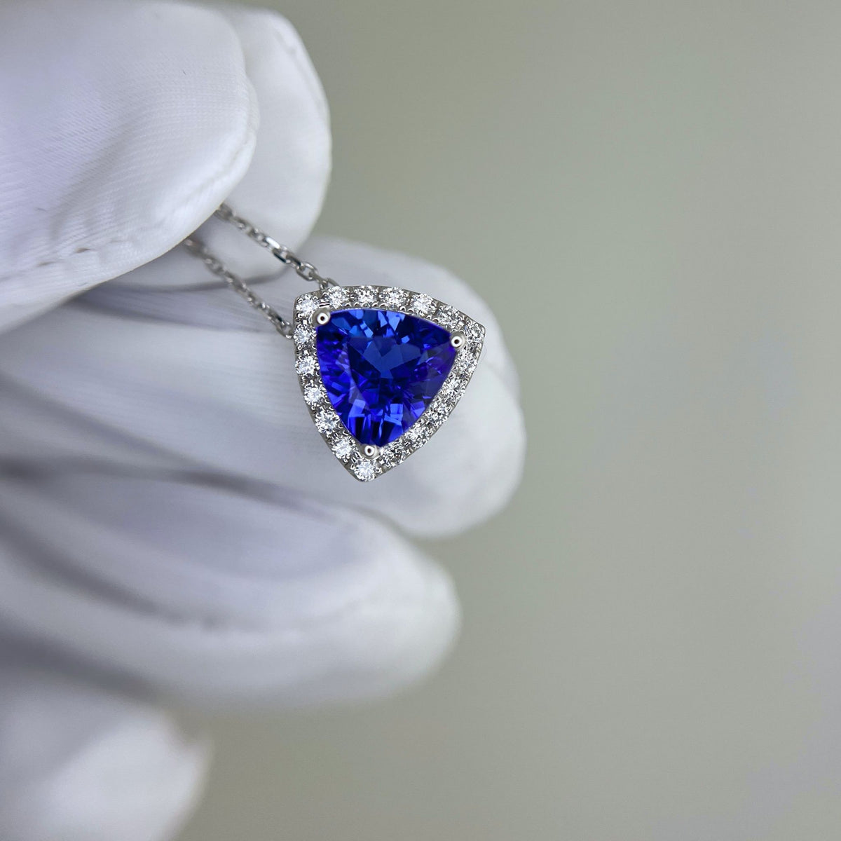 Trillion-cut blue tanzanite gemstone pendant held by a gloved hand against a neutral background