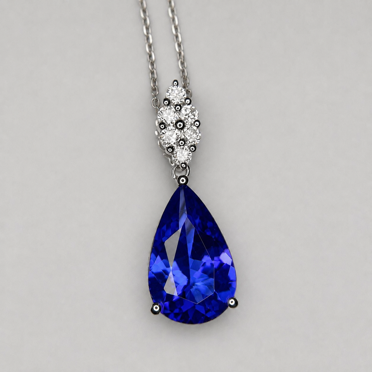 A teardrop-shaped blue gemstone pendant necklace with a silver-toned chain and a decorative silver-toned bail accented with small clear stones, shown on a neutral light gray background.